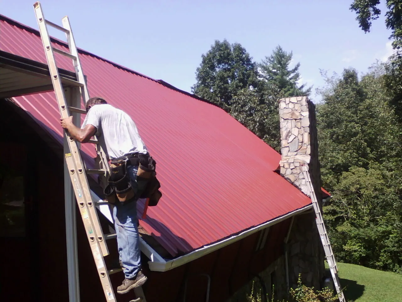 Roofer on a ladder beside a red metal roof and stone chimney — Soffit & Fascia Repair by Roofing Brothers in Markleville